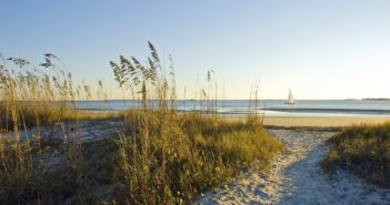 A sand pathway leads to the beach with a sailboat in the background on Hilton Head Island, SC.