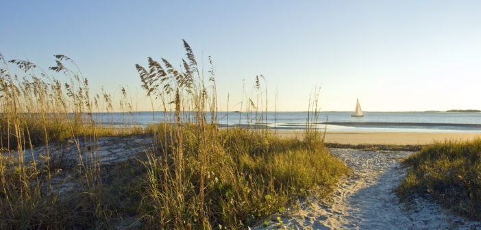 A sand pathway leads to the beach with a sailboat in the background on Hilton Head Island, SC.